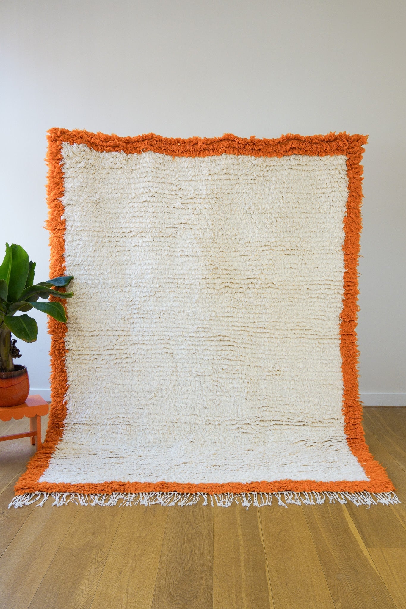 Full view of the Aisha handmade Berber rug with orange border and cream wool, upright on wooden floor