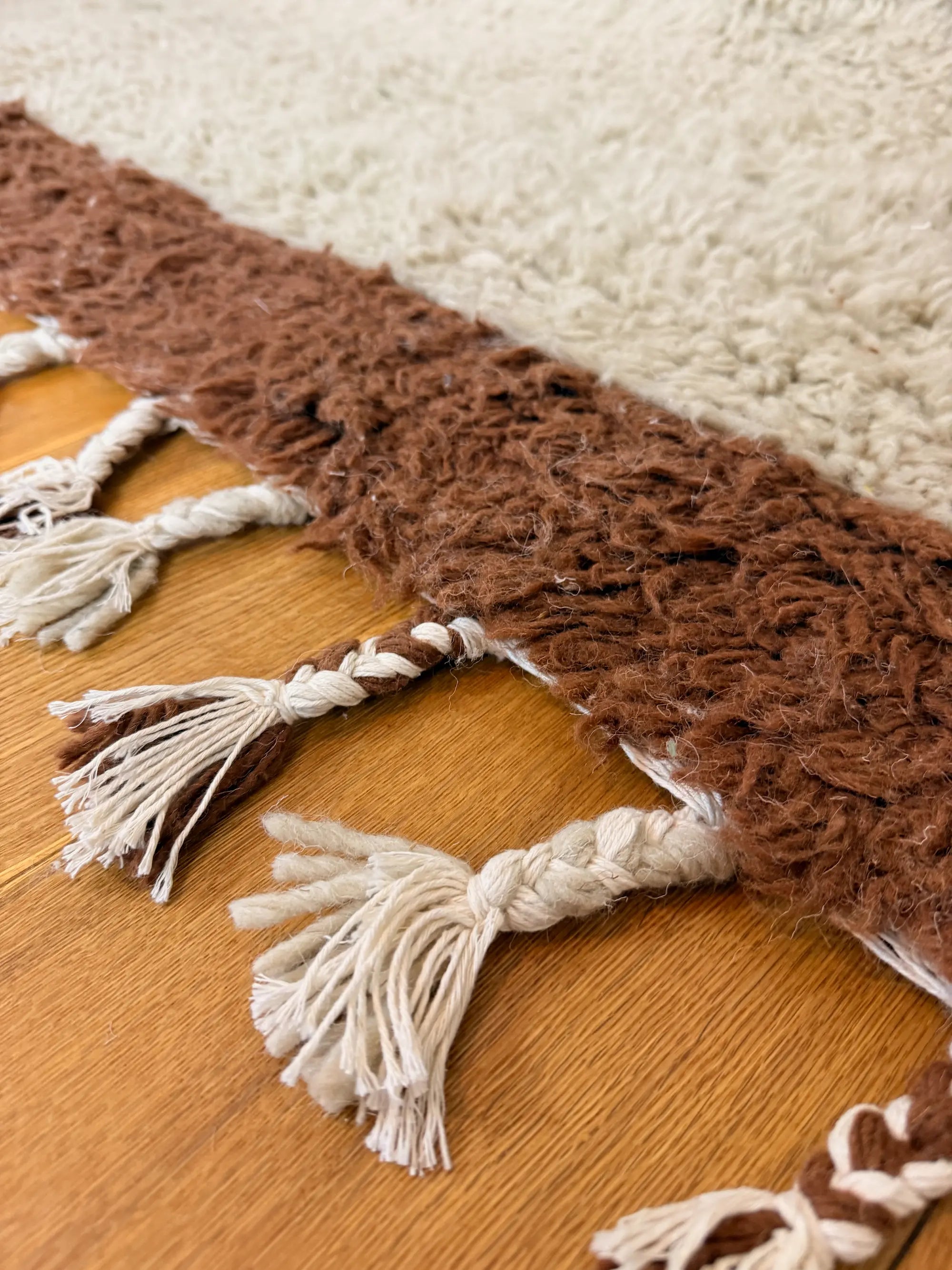 Close-up of the hand-knotted wool fringe ends on the Aliyah shaggy Berber rug, natural white tassels against the chocolate brown border, handmade in Tunisia.
