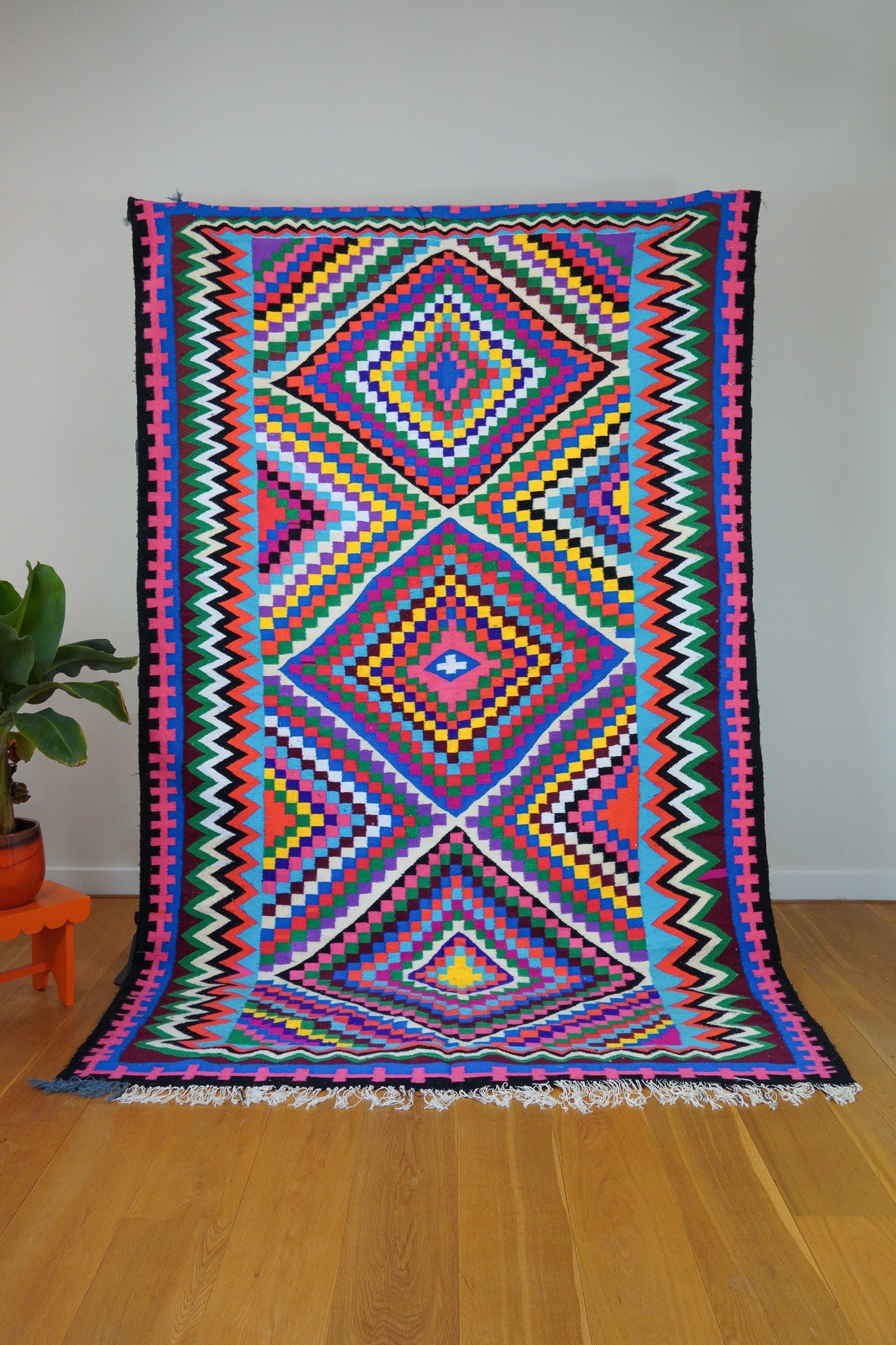 A multicolored geometric checkered patterned handwoven wool rug displayed on a wooden floor against a white wall, with a small orange table and plant to the side.