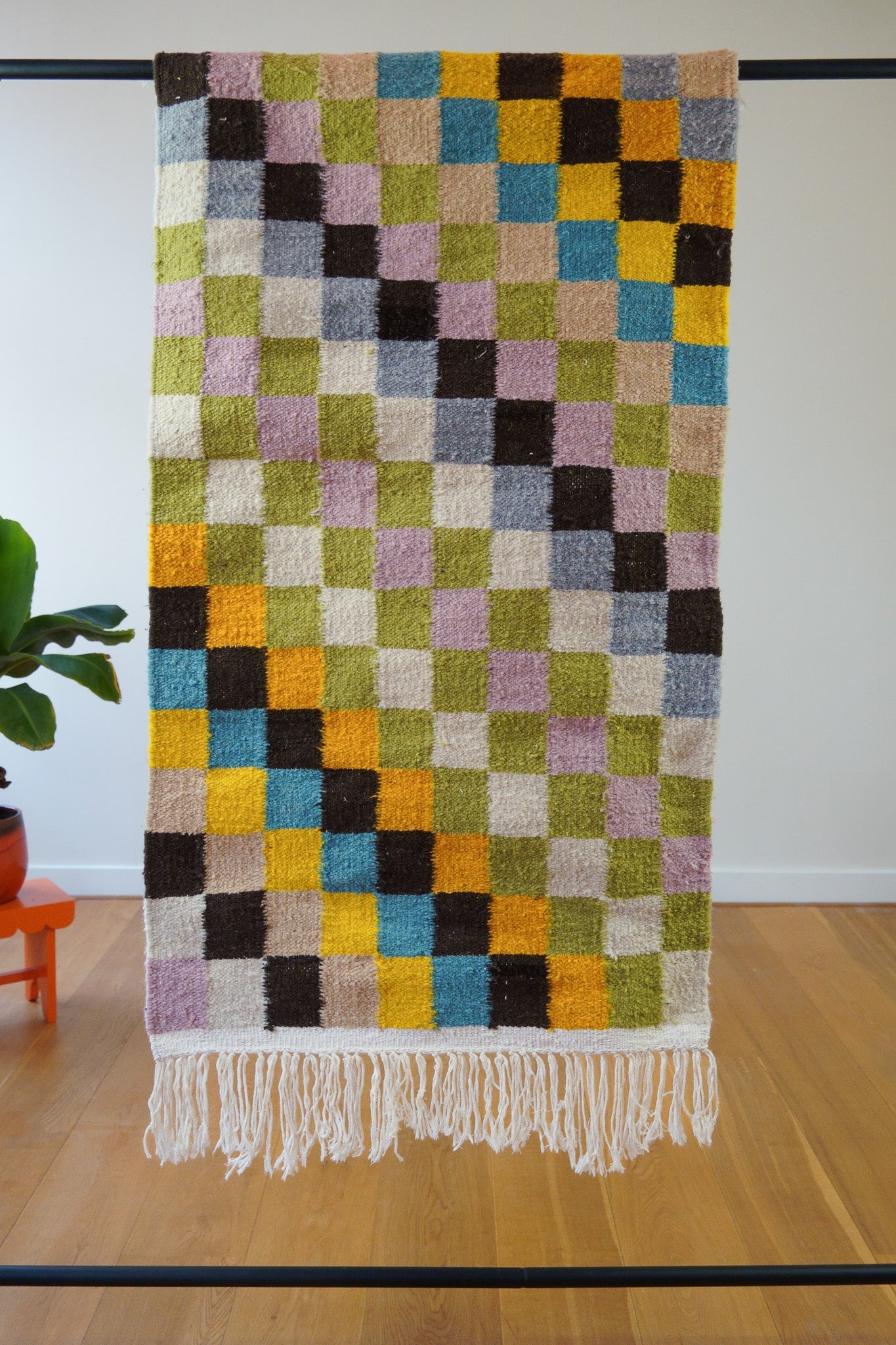 Full view of the Hiba handmade coloured checkered wool rug upright against a white wall, showing vibrant off-white, pink, blue, and yellow hues