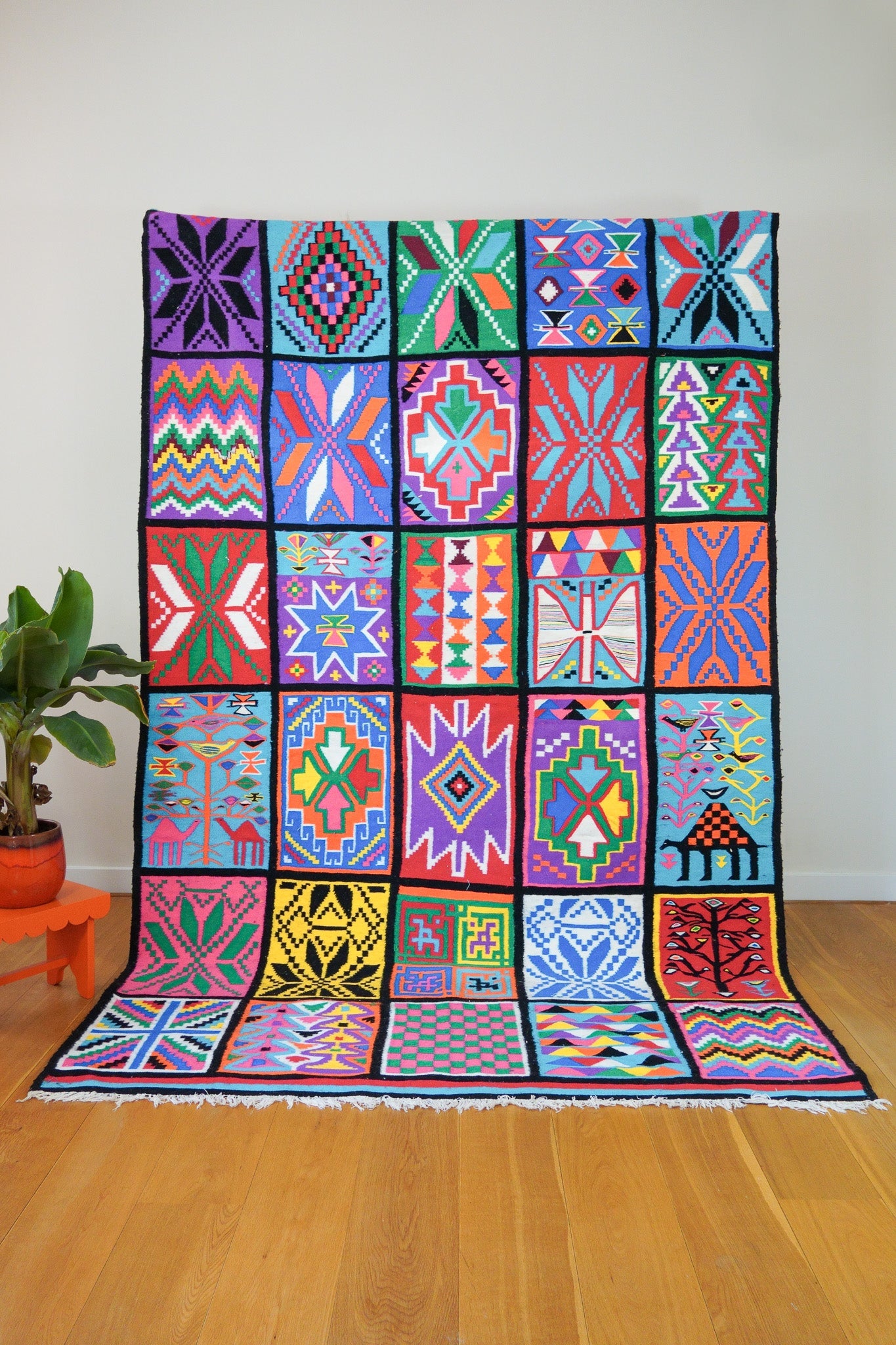 A handwoven colorful wool rug displayed on a wall, featuring a geometric patchwork pattern with multiple colors.