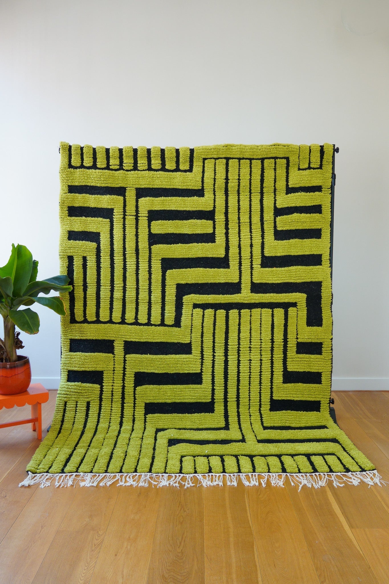 A handwoven Kenza wool rug with a bold geometric design pattern in green and black, displayed on a wooden floor against a white wall – full front view