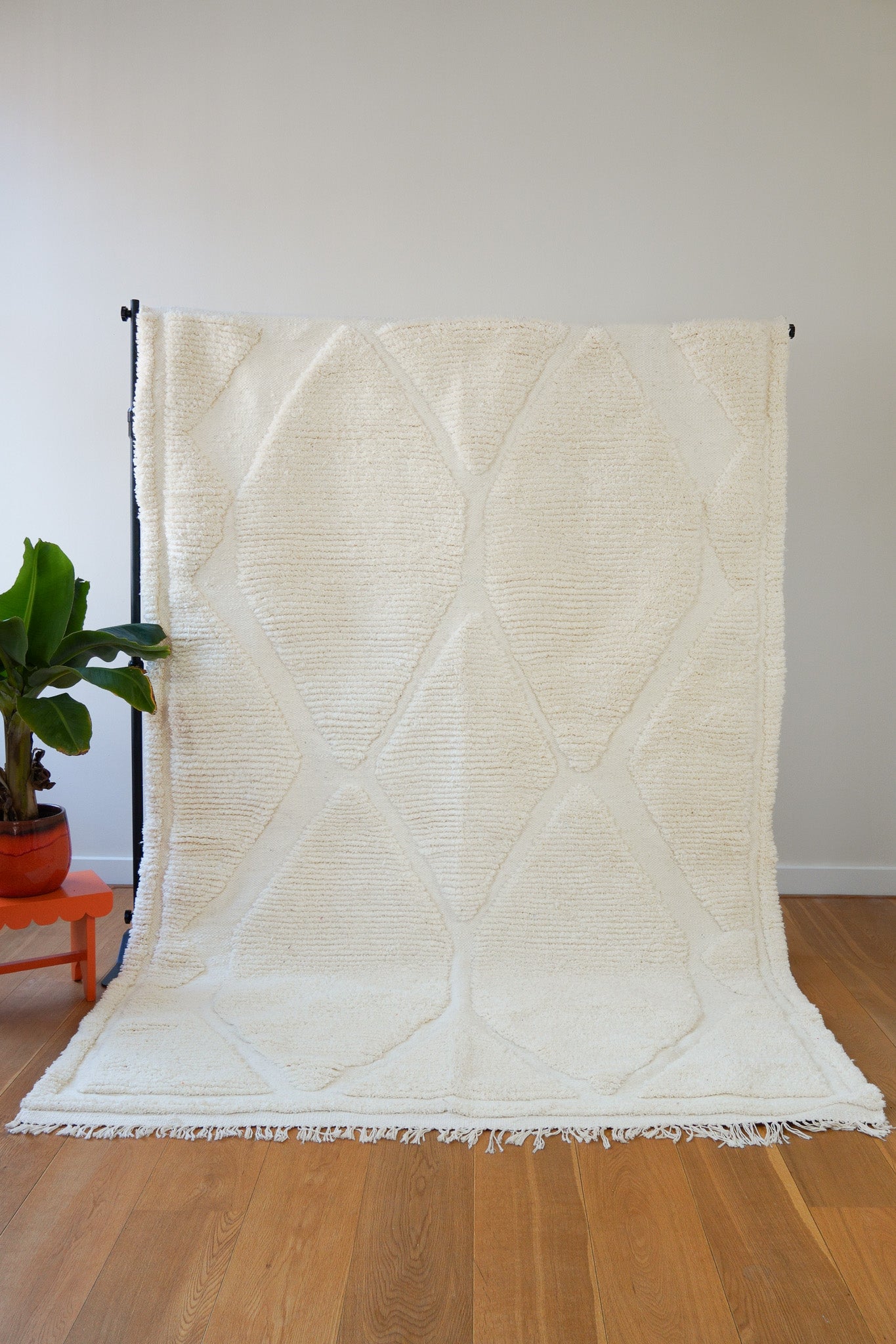 A handwoven ivory wool rug with subtle diamond patterns displayed on a wooden floor against a white wall, with a plant to the side - front view