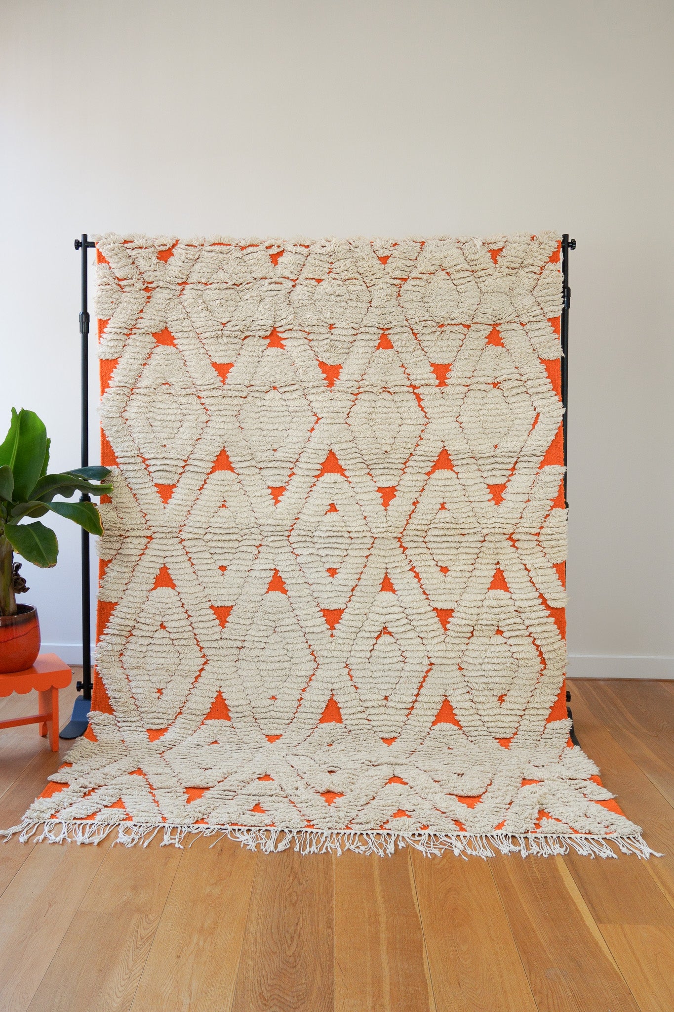 A handwoven orange and cream wool rug with geometric diamond patterns displayed on a wooden floor against a white wall, with a plant to the side, full front view.