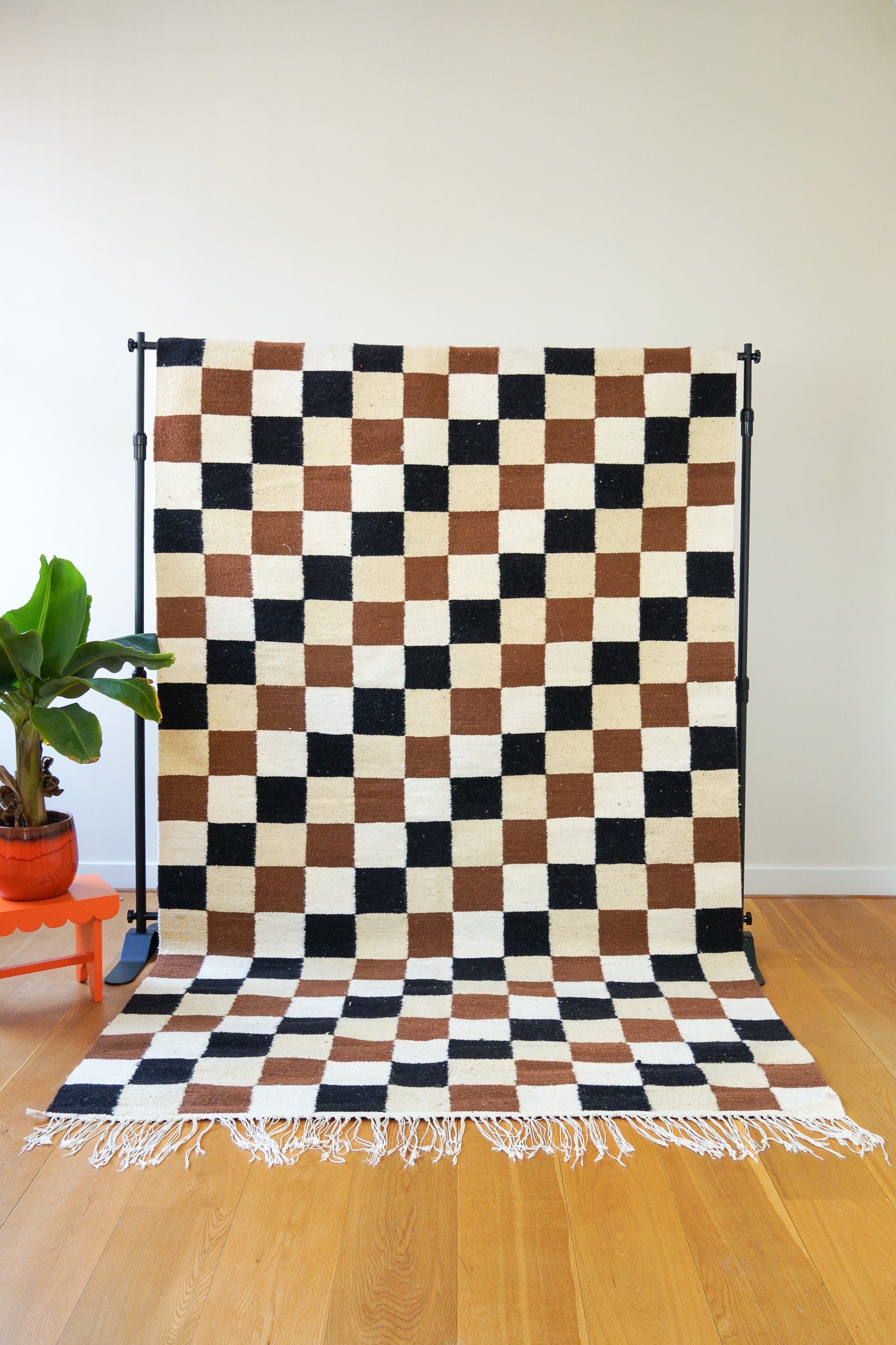 A handwoven wool rug with a geometric checkered design in black, off-white, and brown displayed on a wooden floor against a white wall, with a plant to the side.