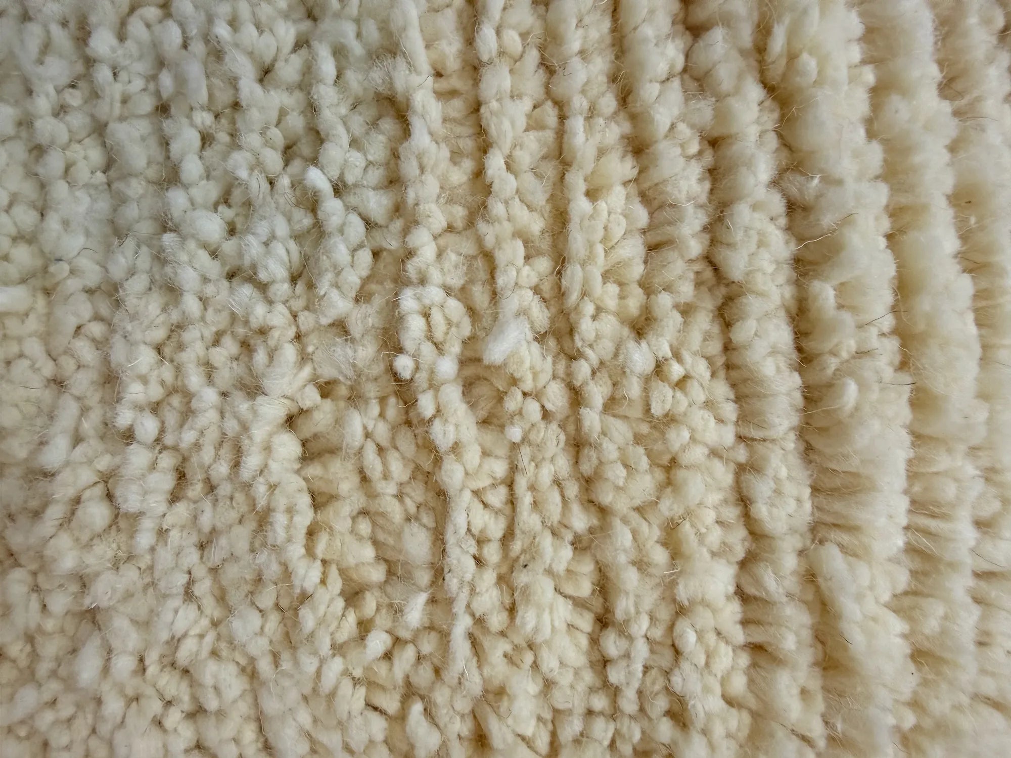 Macro close-up of the natural ivory wool pile on the Beya Berber rug, showing the dense hand-knotted texture and tonal line relief, handmade in Tunisia.